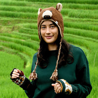 A woman wearing the Hand Crochet Grizzly Hat with sherpa lining, along with a green sweater and fingerless gloves, stands outdoors in front of terraced green fields, looking at the camera and smiling slightly.