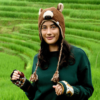 A woman wearing the Hand Crochet Grizzly Hat with sherpa lining, along with a green sweater and fingerless gloves, stands outdoors in front of terraced green fields, looking at the camera and smiling slightly.