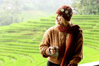 Wearing the Hand Knit Bloom Hat, a person in a brown sweater and scarf stands before lush green rice terraces, holding a glass mug. Light rain falls as they look to the right with a slight smile.