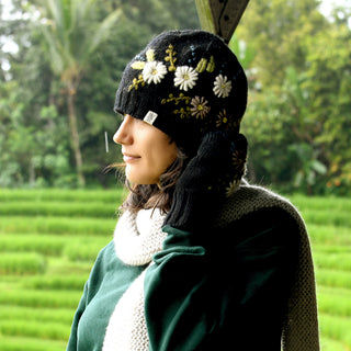 Wearing the Hand Knit Bloom Hat—a black, hand-knit wool piece adorned with white flowers—a person stands outdoors near green fields, holding a matching glove to their ear and smiling slightly. They pair the hat with a green sweater and a white scarf.
