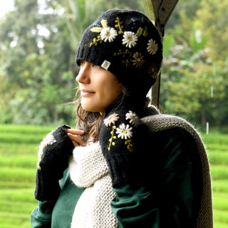 A person wearing the Hand Knit Bloom Hat—a black, hand-knit wool hat featuring white flower embroidery—and matching gloves with a white scarf stands outdoors, looking to the left, with green foliage and a blurred field in the background.