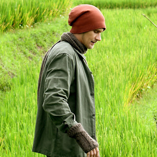 A man wearing the red Dekalb Slouch Merino wool hat, green jacket, and gray scarf stands in a lush green field, gazing down with his left arm slightly extended.