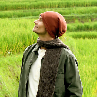 A person wearing the Dekalb Slouch hand-knit red beanie, a dark scarf, and a green jacket looks up while standing in a lush green field with terraced grass in the background.
