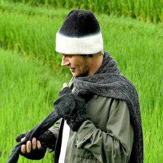 A person wearing the Hand Knit Flow Hat, along with a dark scarf and gloves, stands outside in front of tall green grass, looking down and adjusting their scarf with both hands.