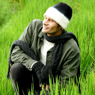 Wearing the Hand Knit Flow Hat—a black-and-white 100% wool beanie—along with a green jacket, white shirt, black scarf, and gloves, a person crouches in tall green grass, looking to the side and smiling.