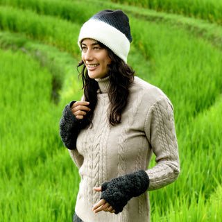 A person wearing a beige cable-knit sweater, fingerless gloves, and the Hand Knit Flow Hat—a black and white 100% wool beanie—stands outdoors in a green field, smiling and looking to the side. The background features lush, terraced grass.