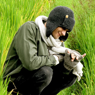 A person crouches in tall green grass, wearing a green jacket, a chunky gray scarf, gray fingerless gloves, and the Elk Hat—a dark wool hat with ear flaps handmade in Nepal—looking down and gesturing with their right hand.