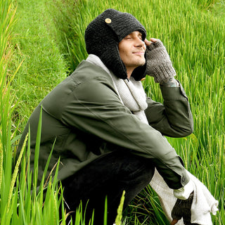 Wearing the Elk Hat, a person smiles with eyes closed as they squat in a grassy field surrounded by tall green plants, dressed in a green jacket, gray scarf, and faux shearling gloves.