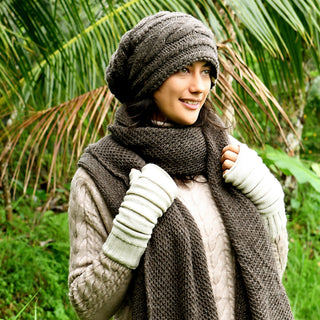 Wearing the Triple Braid Cable Slouch grey Nepal hand-knit beanie, plus a matching scarf and fingerless gloves with a cream cable knit sweater, someone stands outdoors before lush green tropical plants.