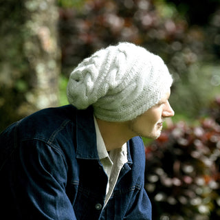 A person wearing the Triple Braid Cable Slouch, a white hand-knit beanie made in Nepal, and a denim jacket is sitting outdoors, looking to the right. The background is blurred with greenery and some brown foliage visible.