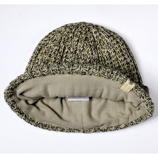 The Hand Knit Grid hat is a handmade beanie in grey and black tones with a folded brim and soft beige fleece lining, shown from above on a white background. This fleece lined beanie features a small fabric tag attached to the brim for a subtle finishing touch.