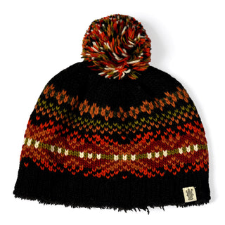 The Hand Knit Explore Hat is a Fair Isle beanie showcasing a multicolor pattern in orange, red, green, and cream on a black background. This knitted hat is topped with a coordinating multicolored pompom and finished with a small fabric tag at the bottom right.