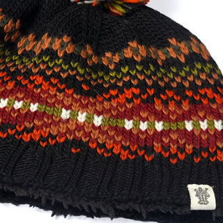 The Hand Knit Explore Hat showcases a bold Fair Isle design with geometric patterns in orange, red, olive green, and white on a black backdrop. This wool beanie is hand-knit and finished with a ribbed band at the bottom and features a cloth tag sewn onto the edge.