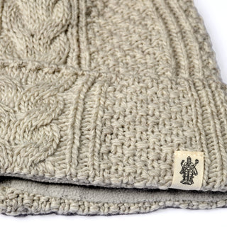 Close-up of the Hand Knit Inter hat in beige, showcasing its classic cable knit pattern and a small corner label featuring a black illustration of a multi-armed figure. The thick, textured fabric suggests this beanie is hand-knit from wool.