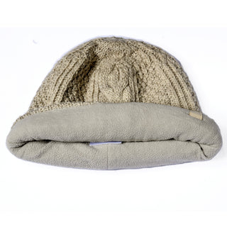 A Hand Knit Inter hat in beige is displayed on a white background. This cable knit beanie features an intricate hand-knit pattern, a large folded fleece brim, and a soft plush lining for added warmth and comfort.