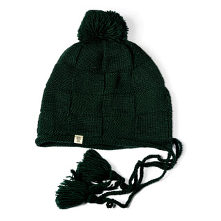 The Hand Knit Nest Hat is a dark green, sherpa-lined earflap hat featuring a pom-pom on top, cozy ear flaps, and two braided tassels with tassel ends. This handmade Nepal hat is finished with a small rectangular metal tag attached to the front edge for a stylish touch.