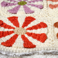Close-up of the Hand Crochet Petal Hood showcasing intricate floral patterns with red and purple petals on a cream background, reminiscent of delicate crocheted daisies. The detailed stitching and textured yarn are clearly visible.