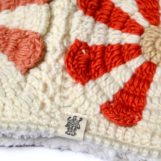 Close-up of the Hand Crochet Petal Hood, showcasing its cream-colored, textured crochet with red and pink floral-inspired patterns reminiscent of daisies, and featuring a small fabric tag with a black line drawing of a figure holding an object.