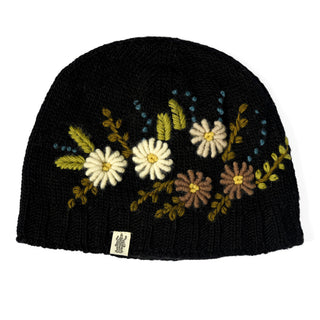 The Hand Knit Bloom Hat is a black knit beanie adorned with embroidered flowers and leaves in white, yellow, green, and brown. Reminiscent of a handmade flower hat, it also features a small tag with a logo sewn onto the bottom edge for an extra touch of detail.