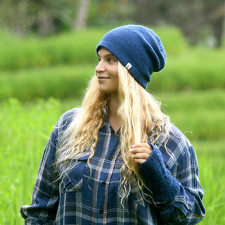 A person with long, wavy blond hair wears The Depp Slouch—a blue slouchy beanie—with a blue plaid shirt, standing outdoors on green grass and looking to the side.
