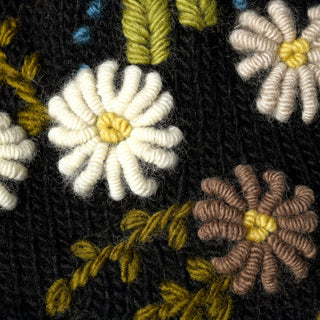 Close-up of embroidered flowers in white, yellow, brown, and beige thread on black fabric, with detailed stitching and green stems. The texture of the yarn and fabric is clearly visible, highlighting the handcrafted quality of the Hand Knit Bloom Hat.