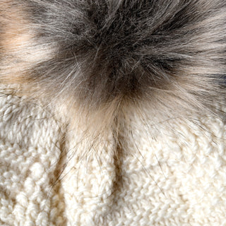 A close-up of a fuzzy gray faux fur pom-pom attached to the Hand Knit Sena Hat, showcasing the detailed texture of its knitted cream-colored yarn and soft fur accent.
