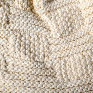 Close-up view of the Hand Knit Sena Hat in a cream color, featuring thick yarn and a textured, repeating pattern. The detailed knitwork showcases various stitches typical of hand knitting, with visible fibers and shadows enhancing the hat’s intricate design.