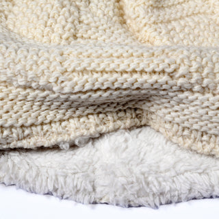 A close-up view of the Hand Knit Sena Hat in cream, layered over a soft, white, fleece-like material, highlights the textured details of both textiles.