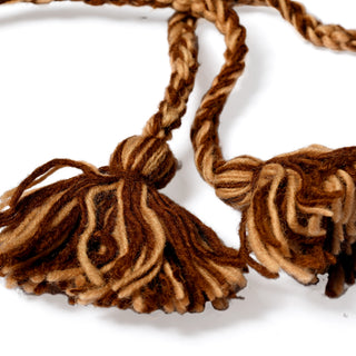Two strands of twisted brown and tan, hand-crocheted wool yarn with large tasseled ends from the Hand Crochet Grizzly Hat lay on a white surface.