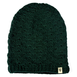 The Hand Knit Puff Beanie, shown in dark green with a textured pattern, is displayed against a white background. A small beige fabric tag featuring a printed design is attached near the bottom edge.