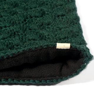 A close-up of a dark green crocheted fabric with a black inner lining, featuring an off-white fabric tag sewn into the edge. The textured material and precise stitching showcase the quality craftsmanship of the Hand Knit Puff Beanie, with details reminiscent of expert hand knitting. The image is well-lit, highlighting the cozy and refined finish that makes this piece perfect for stylish slouchy beanie designs.