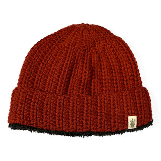 The Hand Crochet Chunky Hat is a red, ribbed knit beanie with a folded brim, featuring a small beige fabric tag with a black design. The inside lining at the bottom edge is black and fuzzy, providing extra warmth and comfort for chilly days.