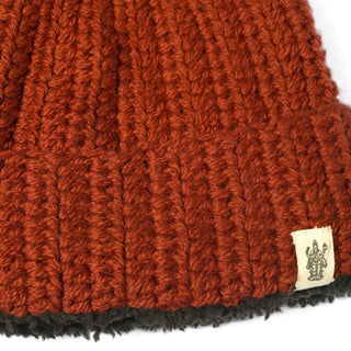 Close-up of the Hand Crochet Chunky Hat in a thick, ribbed, rust-colored lambswool with a small beige tag featuring an illustration of two figures. The lower edge of this cozy winter cap reveals a visible black lining.