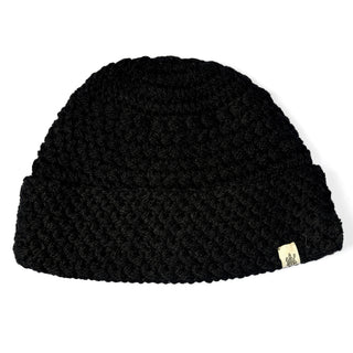 The Hand Knit Hush Hat is a black beanie crafted from lambswool, showcasing a thick, textured hand-knit pattern with a folded cuff. A small beige logo tag is sewn onto the edge of the brim. The beanie is displayed against a white background.
