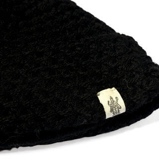 A close-up of a black knitted fabric from the Hand Knit Hush Hat, showing a small white tag with a dark emblem. The knit texture is clearly visible and the tag is sewn near the edge, suggesting it may be part of a folded cuff on this lambswool beanie.