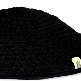 A close-up photo of the Hand Knit Hush Hat—a black crocheted beanie featuring a neatly folded cuff and a small white fabric tag with a simple graphic and text sewn onto the bottom edge—shown against a white background.
