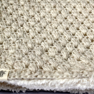 Close-up view of the Hand Crochet Cloud Hood in textured beige, showcasing its visible woven pattern and a small fabric tag at the corner. The material appears thick with a slightly fluffy edge, echoing the look of a hand-crocheted finish.