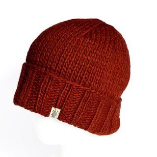 The Hand Knit Timber Hat is a rust-colored, thick knit docker-style beanie featuring a folded brim and a small rectangular tag on the cuff, shown displayed on a white mannequin head against a white background.