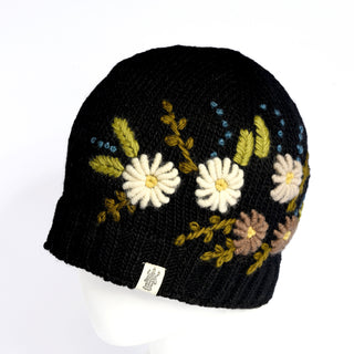 The Hand Knit Bloom Hat is a black knit beanie adorned with embroidered white, yellow, and brown flowers and green leaves, shown on a white mannequin head. This handmade flower hat features a small white tag with a logo attached near the bottom edge.