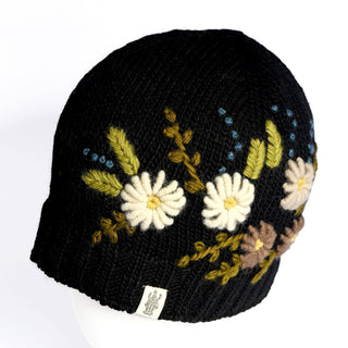 Introducing the Hand Knit Bloom Hat—a black, handmade winter hat crafted from wool, featuring beautifully embroidered white, yellow, blue, and brown flowers with green leaves. This fleece-lined hat is finished with a small white fabric tag bearing a logo near the edge and is displayed on a white mannequin head.