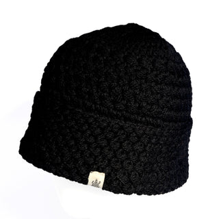 The Hand Knit Hush Hat is a black, hand-crocheted beanie with a textured pattern and a cozy folded cuff. It features a small rectangular fabric tag on the lower edge and is shown against a white background.