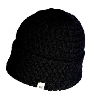 The Hand Knit Hush Hat is a black, crocheted beanie with a subtle textured pattern and a small white tag on the bottom edge. It features a folded cuff detail and is displayed on a white background.