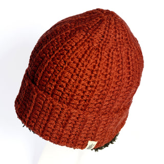 A close-up of the Hand Crochet Chunky Hat in red lambswool is displayed on a white background. This cozy winter beanie features a ribbed pattern and a small beige tag near the edge, shown from a side angle.