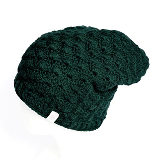 The Hand Knit Puff Beanie in dark green, crafted from Merino wool with a textured pattern and finished with a small white fabric tag on the edge, is displayed on a white mannequin head and shown from the side.