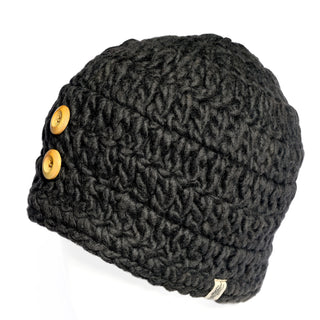 The Hand Crochet Acorn Hat is a black, thickly knit beanie adorned with two large wooden buttons on the side, shown here on a white mannequin head. Crafted from soft wool, this cozy winter accessory blends comfort and style, making it the perfect choice for chilly days.
