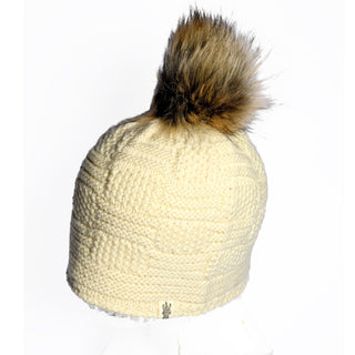 The Hand Knit Sena Hat is a cream knit winter beanie featuring a cozy textured pattern and topped with a large brown faux fur pom-pom. A small rectangular tag accents the bottom edge, and the hat is displayed against a plain white background.