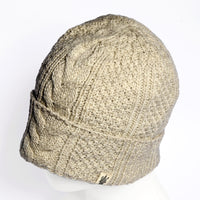 The Hand Knit Inter hat, a beige cable knit beanie crafted from wool, is displayed on a white mannequin head. This hand knit hat features a textured pattern and is finished with a small rectangular tag with a dark design sewn near the edge, providing both stylish flair and cozy warmth.