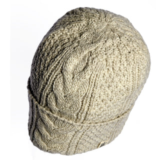 The Hand Knit Inter hat is a beige wool beanie featuring an intricate cable knit pattern and a folded brim, shown against a white background. This hand-knit hat blends classic style with cozy warmth for chilly days.