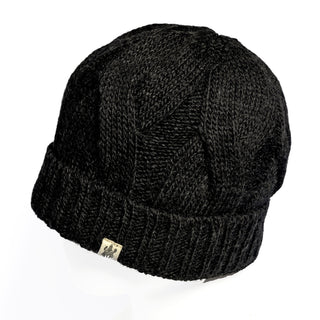The Hand Knit Nord Hat is a black cuffed beanie showcasing a cable pattern and a ribbed folded brim. Made from hand knit wool, it’s accented with a small beige fabric tag featuring a logo on the brim. The hat is presented against a white background.