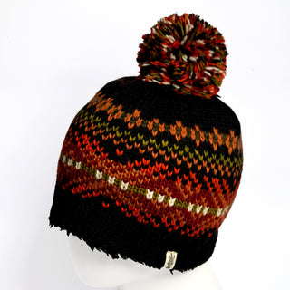 The Hand Knit Explore Hat is a cozy Fair Isle beanie, crafted from wool with a pom-pom on top. It features a black base adorned with multicolored patterns in red, orange, green, and beige, and is shown displayed on a white mannequin head.